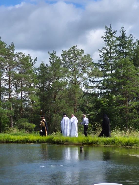 Clergy and parishioners in white at the edge of a pond ringed by pines under a summer sky