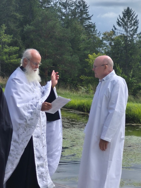 A priest with a white beard reading the prayers of baptism beside the water