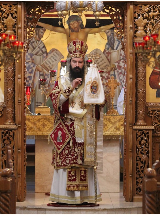 The bishop in red and gold vestments and mitre, holding his staff, standing in the royal doors before the altar and an icon of the Crucifixion