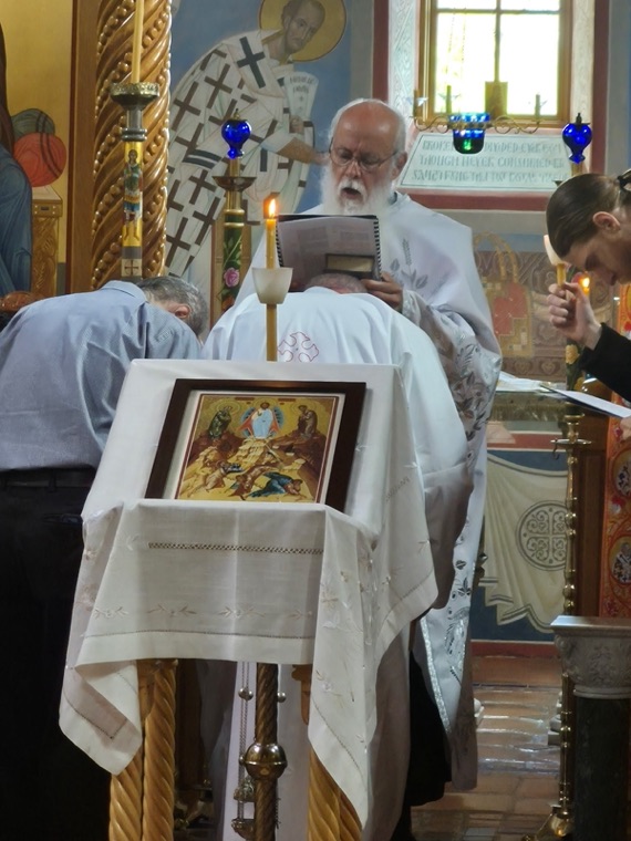 A priest reading prayers over a kneeling man, with a framed icon resting on a cloth-covered stand