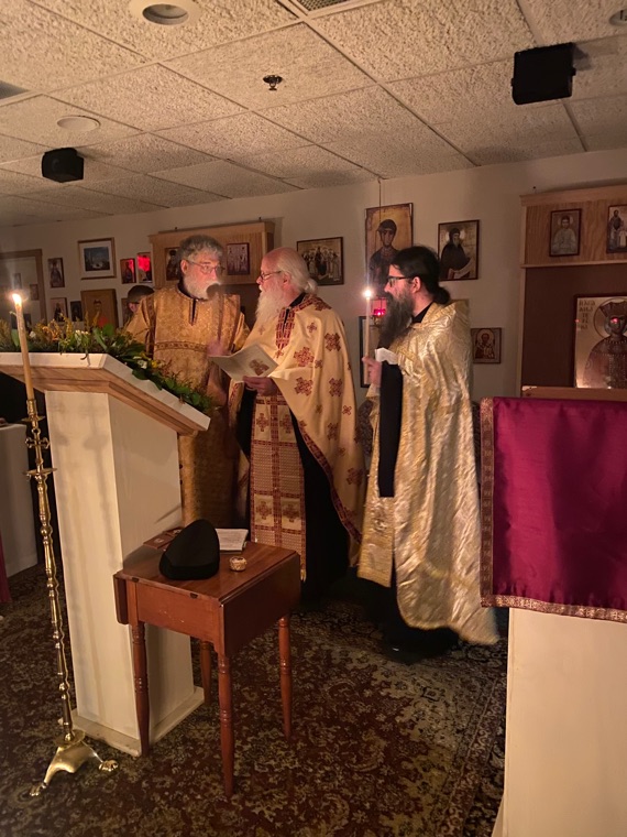 Three clergy in gold vestments reading prayers by candlelight beside a lectern decked with greenery