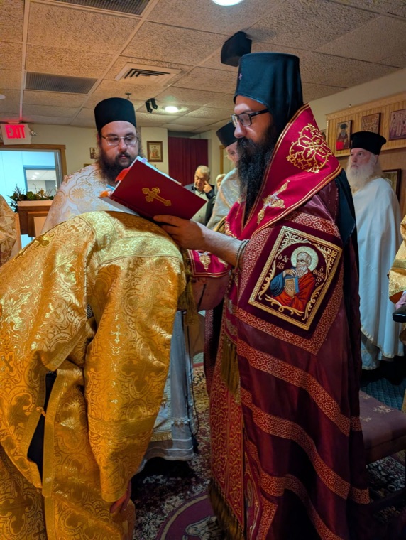 The bishop in red vestments holding a red service book over the head of a man bowing in gold vestments