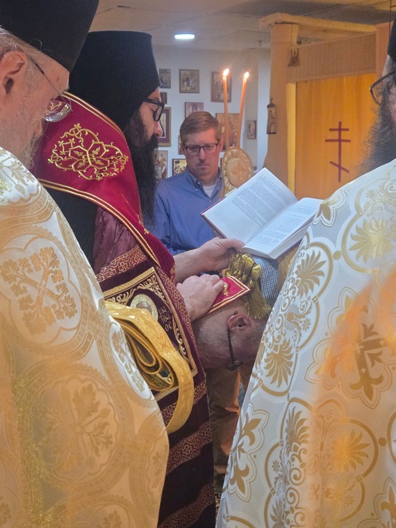 The bishop reading prayers with his hand on the bowed head of a man, surrounded by clergy in white and gold