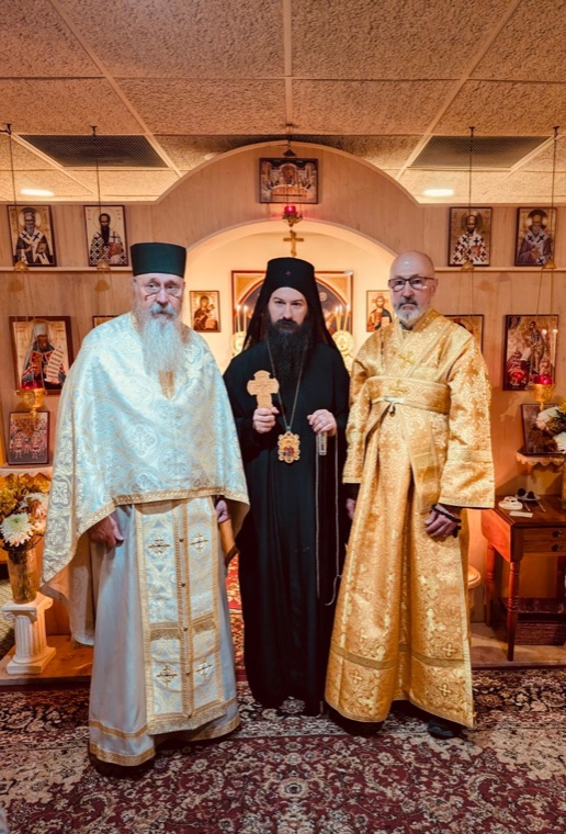 The bishop in black with a hand cross, standing between a priest in white vestments and a deacon in gold before the iconostasis