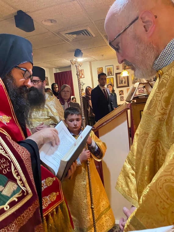 The bishop reading from a service book to a deacon in gold vestments, while an altar boy holds the book and the parish looks on